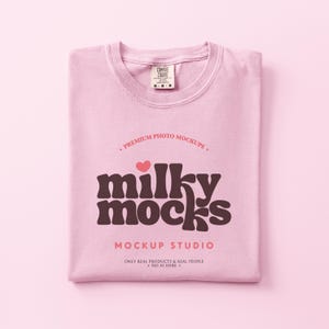 May include: A folded light pink t-shirt with the text "milky mocks" in a brown retro font, a small pink heart, and the words "MOCKUP STUDIO". The shirt has a Comfort Colors tag.