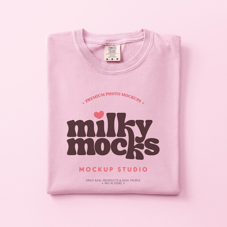 May include: A folded light pink t-shirt with the text "milky mocks" in a brown retro font, a small pink heart, and the words "MOCKUP STUDIO". The shirt has a Comfort Colors tag.