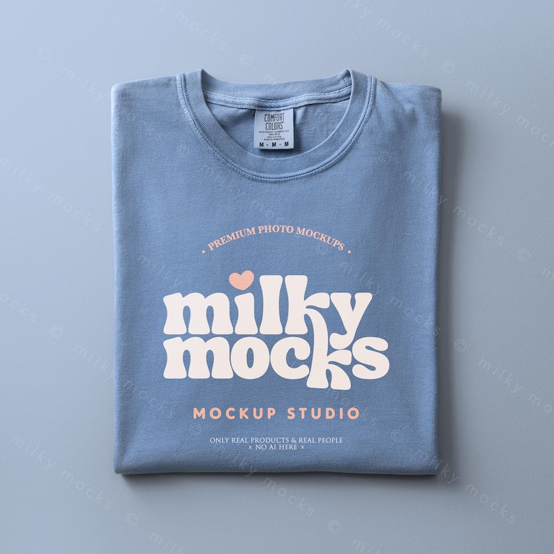 May include: A folded, light blue t-shirt with the text "milky mocks" in a retro font, a heart graphic, and the words "Mockup Studio". The shirt has a tag that says "Comfort Colors". The shirt is a mockup.