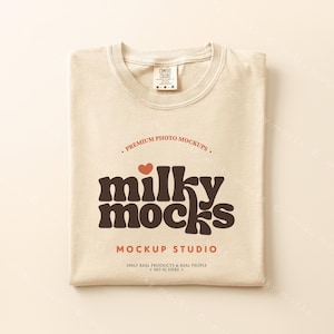 May include: A folded, cream-colored t-shirt with the text "milky mocks" in brown, a red heart, and the words "MOCKUP STUDIO". The shirt has a tag that says "COMFORT COLORS". The shirt is folded neatly on a light beige surface.