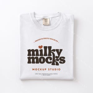 May include: A folded white t-shirt with the text "milky mocks" in brown, a brown heart, and the words "MOCKUP STUDIO". The shirt has a tag that says "COMFORT COLORS". The shirt is folded neatly.