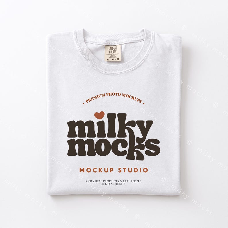 May include: A folded white t-shirt with the text "milky mocks" in brown, a brown heart, and the words "MOCKUP STUDIO". The shirt has a tag that says "COMFORT COLORS". The shirt is folded neatly.