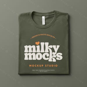 Puede incluir: Camiseta verde oliva doblada, con cuello redondo. La camiseta presenta el texto "milky mocks" en una fuente retro, con un coraz&oacute;n naranja. Tambi&eacute;n son visibles las palabras "MOCKUP STUDIO" y "ONLY REAL PRODUCTS & REAL PEOPLE".