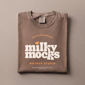 May include: A folded brown t-shirt with the text "milky mocks" in a retro font. The shirt has a crew neck and a tag that reads "Comfort Colors". The shirt is a medium size. The words "Premium Photo Mockups" and "Mockup Studio" are also printed on the shirt.