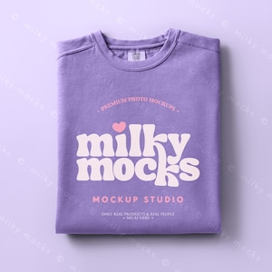 May include: A folded lavender t-shirt with the text "milky mocks" in a retro font, a pink heart, and the words "Mockup Studio". The shirt is a Comfort Colors brand and is on a matching lavender background.