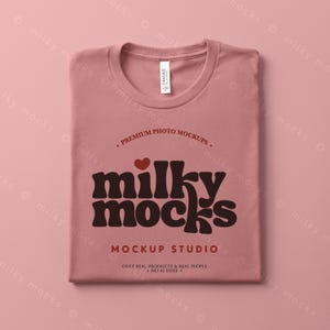 Puede incluir: Una camiseta doblada, de color rosa polvoriento, con cuello redondo. La camiseta presenta el texto "milky mocks" en una fuente retro marr&oacute;n, con un coraz&oacute;n rojo encima de la palabra. El texto adicional incluye "PREMIUM PHOTO MOCKUPS" y "MOCKUP STUDIO".