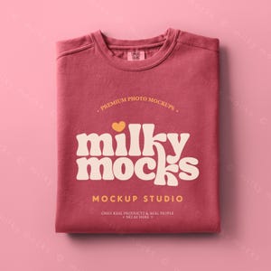 Può includere: Una maglietta piegata, color rosa polveroso, con la scritta "milky mocks" in un font retrò. La maglietta ha uno scollo tondo e si trova su uno sfondo rosa. Il testo "MOCKUP STUDIO" è sotto.