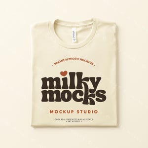 Puede incluir: Una camiseta crema doblada con cuello redondo. La camiseta presenta el texto "milky mocks" en marr&oacute;n, con un coraz&oacute;n rojo encima de la palabra. El texto adicional incluye "Premium Photo Mockups" y "Mockup Studio."