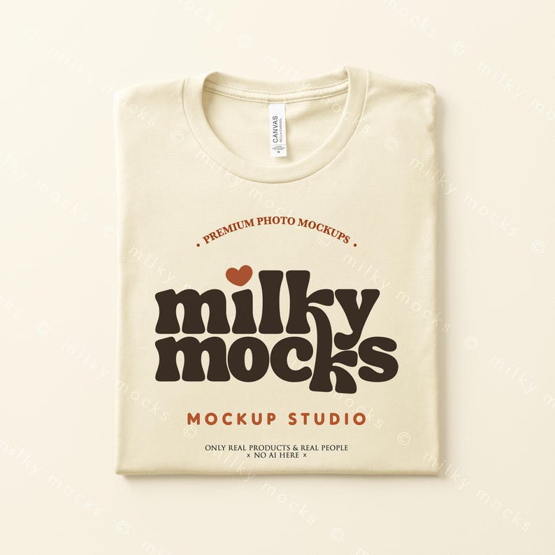 Puede incluir: Una camiseta crema doblada con cuello redondo. La camiseta presenta el texto "milky mocks" en marr&oacute;n, con un coraz&oacute;n rojo encima de la palabra. El texto adicional incluye "Premium Photo Mockups" y "Mockup Studio."