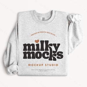 May include: A pale grey sweatshirt featuring the words "milky mocks" in a retro font. The sweatshirt has a crew neck and long sleeves. The text "Premium Photo Mockups" and "Mockup Studio" are also printed on the sweatshirt.