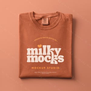 May include: A folded rust-colored t-shirt with the text "milky mocks" in a retro font. The shirt has a small tag at the neckline and the words "MOCKUP STUDIO" are printed below the main text. The shirt is on a matching rust-colored background.