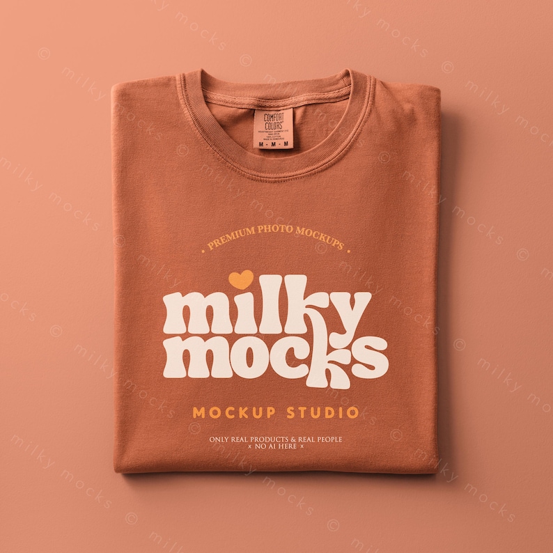 May include: A folded rust-colored t-shirt with the text "milky mocks" in a retro font. The shirt has a small tag at the neckline and the words "MOCKUP STUDIO" are printed below the main text. The shirt is on a matching rust-colored background.
