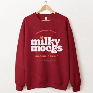 May include: A maroon crewneck sweatshirt with the text "milky mocks" in white, with an orange heart above the text. The sweatshirt is hanging on a wooden hanger. The text "MOCKUP STUDIO" is below the main text.