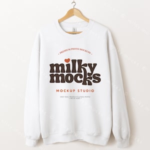 Könnte beinhalten: Ein weißes Sweatshirt mit Rundhalsausschnitt und dem braunen Text "milky mocks", einem roten Herz und den Worten "MOCKUP STUDIO". Das Sweatshirt hängt an einem Holzbügel.