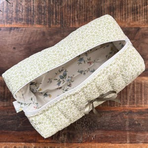 May include: A green quilted makeup bag with a floral print and a brown ribbon tie. The bag is open and the inside is lined with a white fabric with a smaller floral print.