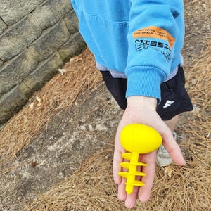 Könnte beinhalten: Ein leuchtend gelbes, eiförmiges Objekt mit einem Stiel und drei gestuften, kreisförmigen Flossen. Das Objekt wird in der Hand einer Person gehalten. Die Person trägt einen blauen Kapuzenpullover mit Text auf dem Ärmel, der lautet: "NATIONAL AERONAUTICS AND SPACE ADMINISTRATION MISSION".