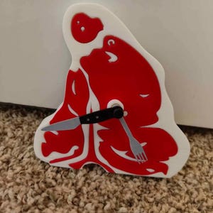 May include: A novelty wall clock shaped like a steak, with a red meat design and a white outline. The clock hands are a knife and fork. The clock is mounted on a white surface.