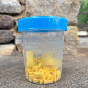 May include: A clear plastic container with a blue lid filled with yellow foam letter shapes. The container has printed text on the side. The container is sitting on a gray surface.