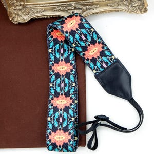 Könnte beinhalten: Ein gemusterter Kameragurt mit schwarzer Basis und einem sich wiederholenden Blumendesign in Koralle, Hellblau und Lila. Der Gurt hat ein schwarzes Lederendstück und eine schwarze verstellbare Kordel.
