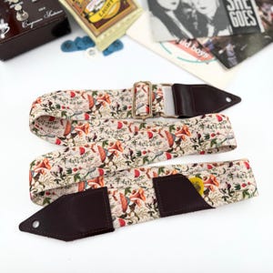 May include: A guitar strap with a cream floral pattern featuring orange, red, and green elements. It has brown leather ends and a gold-coloured metal buckle. A guitar pick is visible in the strap's pocket.