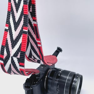 Może przedstawiać: Czarny aparat fotograficzny z paskiem we wzory. Pasek ma wzór w szewrony w kolorze czerwonym, czarnym, białym i różowym. Obiektyw aparatu jest czarny i srebrny, a pasek jest przymocowany czerwonymi skórzanymi końcami.