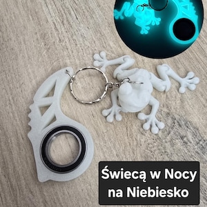 May include: A white frog-shaped keychain with a silver keyring and a black bearing. The keychain has a glowing blue frog design in a circle. The text on the image says "Glows in Blue at Night".