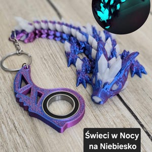 May include: A 3D-printed dragon keychain in shades of purple, blue, and white. The dragon has a detailed, segmented body and a crystal-like structure on its back. A separate, matching keychain component with a bearing is also included. The text "Świeci w Nocy na Niebiesko" is visible.