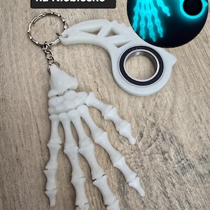 May include: A white, glow-in-the-dark skeleton hand keychain. The keychain has a silver ring and a circular design. The text "Świecą w nocy na Niebiesko" is in the upper left corner. The keychain glows blue in the dark.