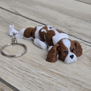 Op de afbeelding: Een bruin en witte Cavalier King Charles Spaniel sleutelhanger. De hond ligt met zijn hoofd op het oppervlak. De sleutelhanger heeft een zilveren metalen ring.