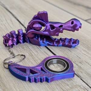 May include: A purple and blue keychain featuring a stylized crocodile skull and a separate, matching piece with a bearing. The keychain is made of a 3D-printed material and includes a silver keyring.