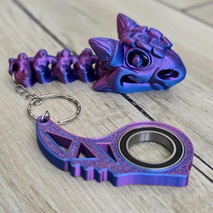 May include: A collection of 3D-printed items in a gradient of purple and blue. Includes a dragon head, a segmented spine, and a key chain with a bearing. The items are on a wooden surface.