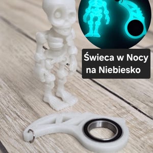 May include: A white skeleton figurine and a white, curved object with a metal bearing are displayed. A glowing blue image of the skeleton is shown in a circle. The text "Świeca w Nocy na Niebiesko" is also present.