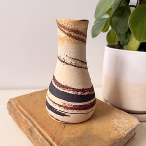 May include: A ceramic vase with a cream-colored base and a series of dark brown, black, and burgundy stripes. The top of the vase is a light brown color. The vase is sitting on top of a vintage book.
