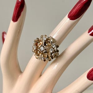 May include: A gold-toned ring featuring a cluster of small, flower-shaped designs. Some of the flowers have clear, round gemstones in the center. The ring is displayed on a mannequin hand with red nail polish.
