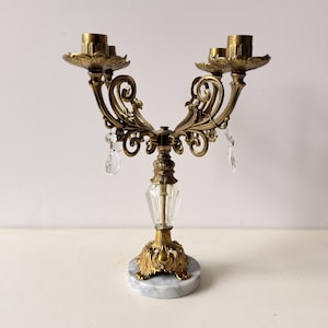 May include: A brass candelabra featuring two candle holders with ornate scrollwork arms, clear crystal teardrop pendants, a central faceted glass stem, and a circular white marble base. The metal components have a weathered gold finish.