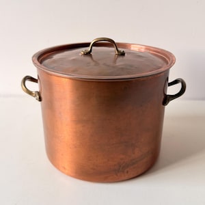 May include: A large, cylindrical copper stockpot with a matching lid. The pot features two curved brass side handles and a small brass handle on the lid. The exterior surface shows a warm, metallic copper finish with subtle signs of wear.