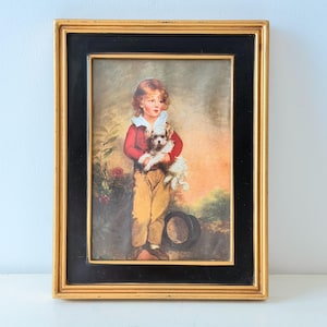 May include: Framed vintage print of a child holding a small dog. The child wears a red jacket, tan pants, and a white collar. The print is in a gold and black frame, with a pastoral background.