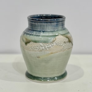 May include: A handcrafted ceramic vase with a unique, textured design. The vase features a gradient of colors, including shades of blue, green, and cream. The textured band adds visual interest to the piece. The vase has a rounded body and a slightly flared rim.