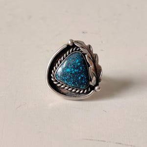 May include: A sterling silver ring featuring a triangular turquoise stone with dark matrix patterns. The stone is framed by a twisted rope border and a decorative leaf motif on one side, set within a polished silver bezel and band structure.