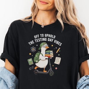 Può includere: T-shirt nero con la scritta "OFF TO UPHOLD THE TESTING DAY VIBES". La maglietta presenta un'oca dei cartoni animati su uno scooter, con occhiali e cappello, che trasporta libri e uno zaino. Grafica aggiuntiva include stelle, una matita e un blocco note.