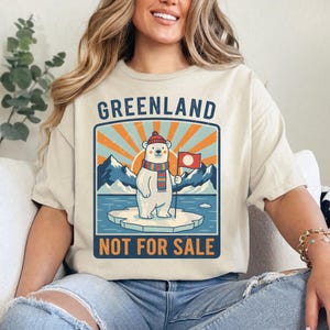 May include: Cream-coloured t-shirt featuring a polar bear graphic on an iceberg, holding a flag. The design includes the word "GREENLAND" above and "NOT FOR SALE" below. The bear wears a hat and scarf, with a mountain and sun background.