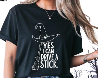 May include: Black t-shirt with a white graphic of a witch's hat and broom. The text "YES I CAN DRIVE A STICK" is printed below the hat. The shirt is a Halloween-themed design.