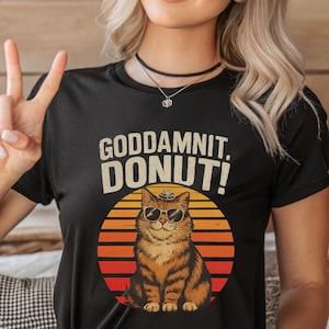 Könnte beinhalten: Schwarzes T-Shirt mit dem Text "GODDAMNIT, DONUT!" über einer Cartoon-Katze mit Sonnenbrille und Hut, vor einem orange-gelben Hintergrund im Sonnenuntergangsstil.