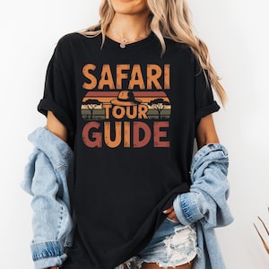 Peut inclure: T-shirt noir avec l'inscription "SAFARI TOUR GUIDE" en orange et marron. Le design comprend un chapeau, des arbres et un fond évoquant un coucher de soleil.