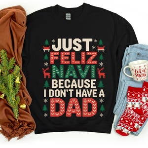 Può includere: Felpa nera con la scritta "JUST FELIZ NAVI BECAUSE I DON'T HAVE A DAD" in bianco, rosso e verde. Il design include alberi di Natale e renne. Sono visibili anche una tazza e calzini.