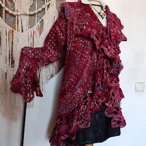May include: A burgundy crochet jacket with long sleeves and a ruffled front. The jacket is displayed on a mannequin, showcasing its intricate openwork design and flowing silhouette. The color palette includes shades of red, blue, and white.