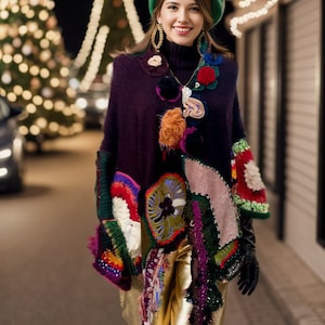 May include: A woman wearing a colorful crocheted poncho with a green beret and gold pants. She is standing in front of a Christmas tree with lights.