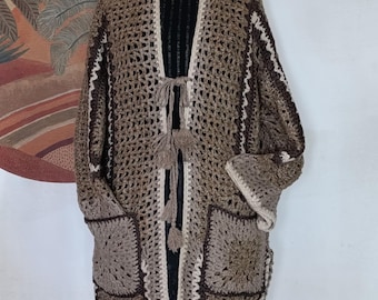Puede incluir: Un cárdigan largo de crochet marrón con cierre de nudo y grandes bolsillos cuadrados. El cárdigan está hecho con una mezcla de hilo marrón y crema.