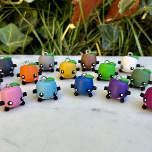May include: A collection of 16 colorful, small, square-shaped charms with a leaf on top. Each charm has a cute face with black eyes and a small mouth. The charms are made of polymer clay and are perfect for adding a touch of whimsy to jewelry or crafts.