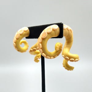 May include: A pair of yellow and white octopus tentacle earrings. The earrings are made of polymer clay and are shaped like curled octopus tentacles with suction cups.